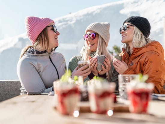Three women sitting on a bench, wearing sunglasses and holding drinks.