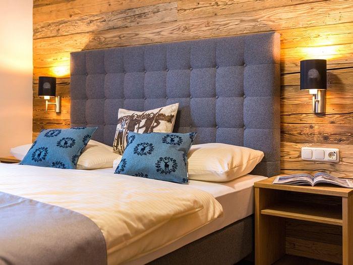 Gemütliches Hotelzimmer mit blauem Polsterkopfteil, rustikaler Holzverkleidung und Zierkissen