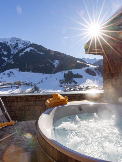 A bubbling hot tub on the terrace with lounge chairs, overlooking a sunny, snow-covered mountain landscape.