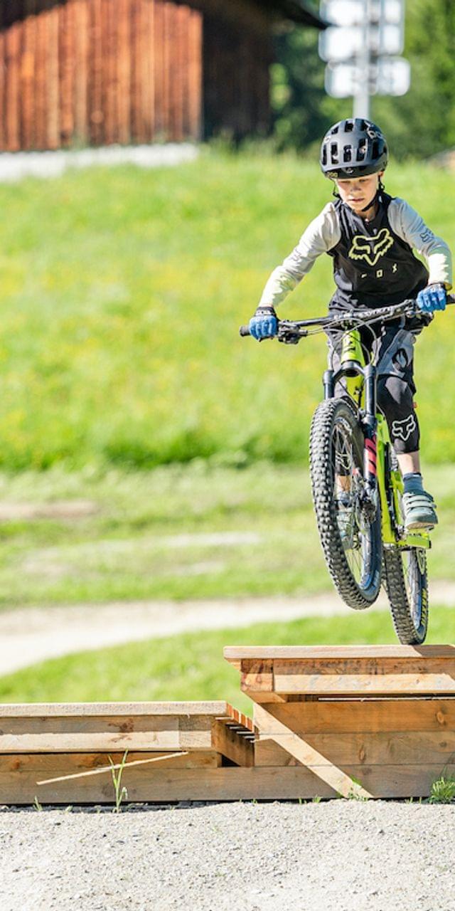 Junge springt mit Mountainbike über Holzrampe, ein anderes Kind schaut in Schutzausrüstung zu