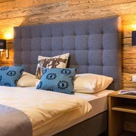 Gemütliches Hotelzimmer mit blauem Polsterkopfteil, rustikaler Holzverkleidung und Zierkissen