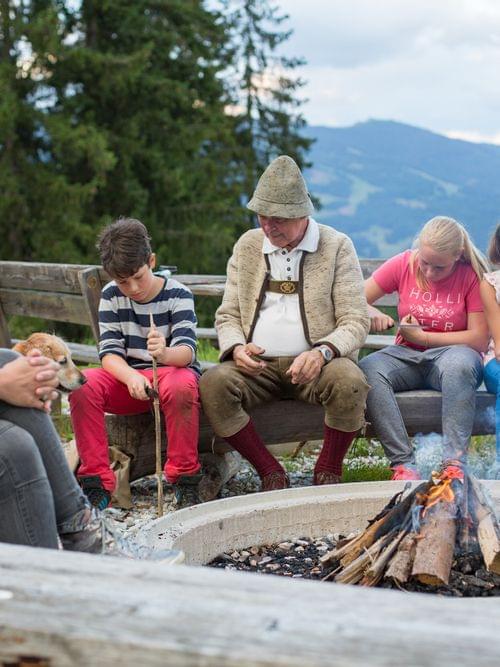Familie und Kinder sitzen am Lagerfeuer mit malerischer Alpenkulisse im Hintergrund