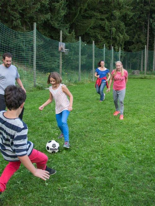 Family playing soccer on a grassy field surrounded by green forest and net fencing