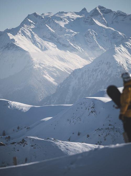 Snowboarder in gelber Jacke blickt auf schneebedecktes Alpengebirge an sonnigem Tag