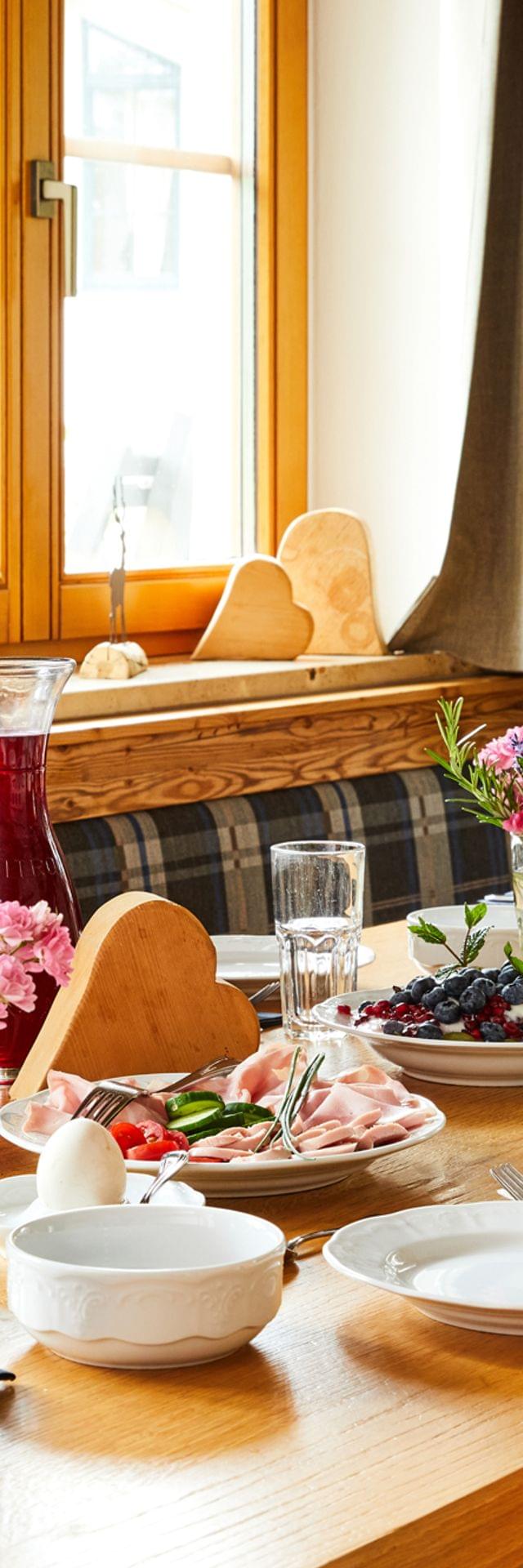 Rustikaler Frühstückstisch mit Brot, Aufschnitt, Beeren, Saft, Kaffee und Blumen am sonnigen Fenster