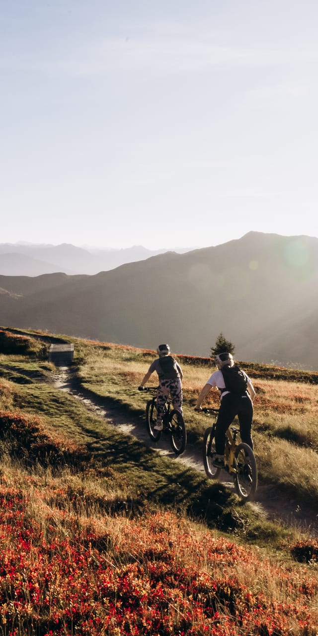 An atmospheric view of the autumn mountain landscape, with two mountain bikers riding down a trail.