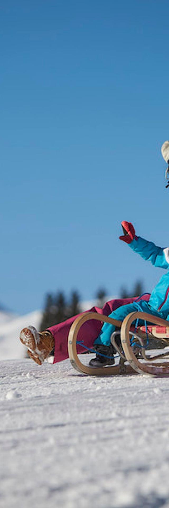 Mutter und Kind rodeln einen verschneiten Hang hinunter vor Alpenkulisse