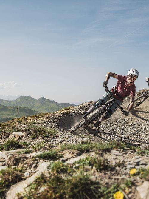 Zwei Mountainbiker fahren eine Kurve auf einem Schotterweg vor alpiner Kulisse