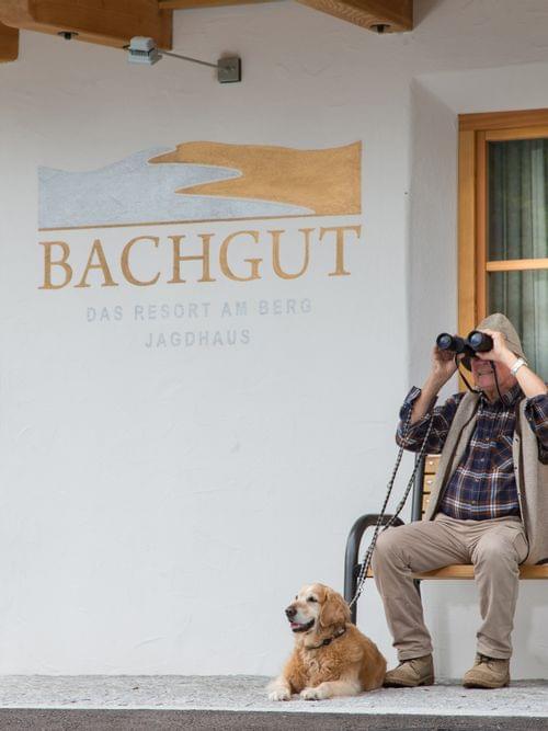 Mann mit Fernglas und Hund auf Bank vor dem Bachgut Bergresort