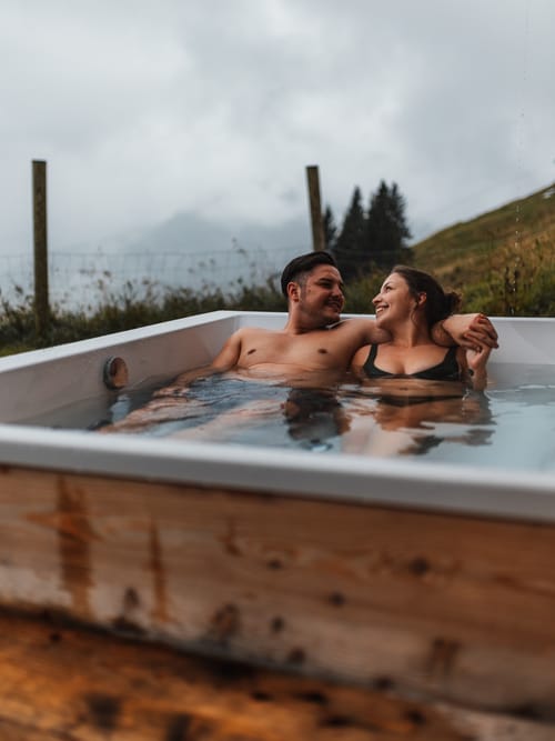 Two people in an outdoor hot tub, with an autumn landscape behind them.