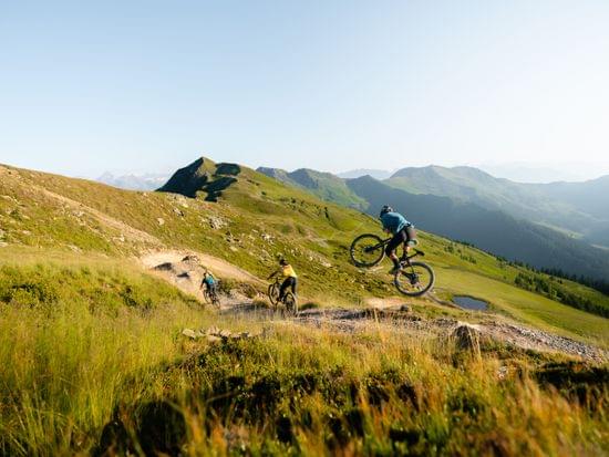 Mountainbiker auf Abfahrtspfad, einer springt in der Luft vor alpiner Berglandschaft