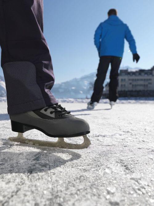 People ice skating outdoors on a frozen lake with snow-covered mountains in the background