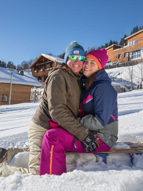 Smiling couple sitting on a sled in snowy Alpine village on sunny winter day