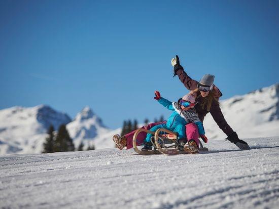 Mother and child sledding down snowy slope with alpine mountains in background