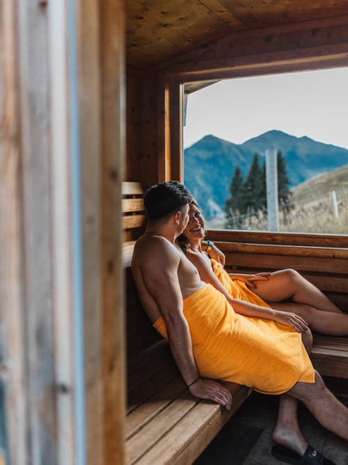 Two people in the sauna, looking out through the panoramic window at the mountain landscape.