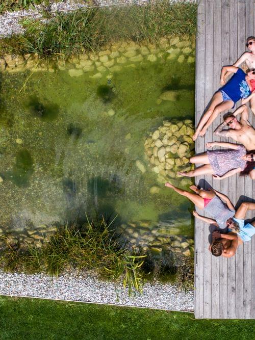Friends relaxing on wooden deck beside natural swimming pond in summer