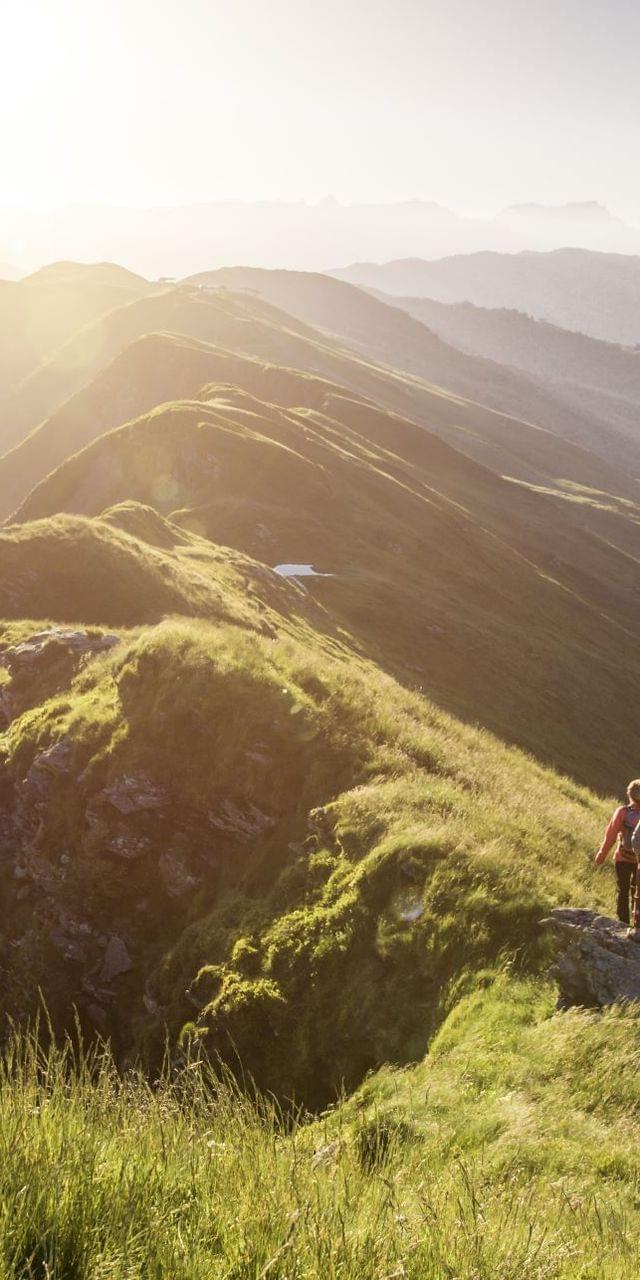Wanderer auf einem Berggrat bei Sonnenaufgang mit alpiner Landschaft im Hintergrund