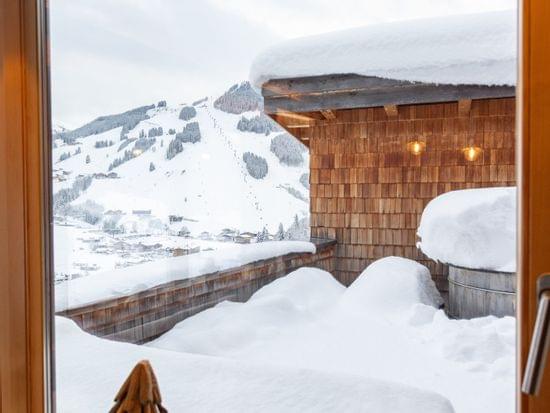 Blick durchs Fenster auf verschneite Chalet-Terrasse und Alpen-Skipisten im Winter
