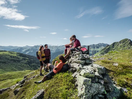 Freunde die am Berg eine Pause machen auf einer Bank, im Hintergrund tief grüne Almwiesen und Sonnenschein 