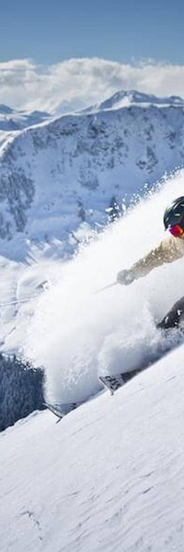 Skifahrer fährt durch frischen Pulverschnee auf steilem Hang mit verschneiten Alpen im Hintergrund