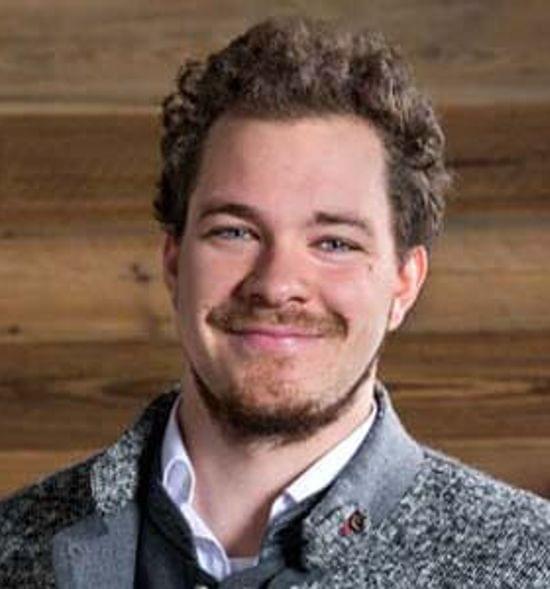 Smiling man in gray blazer with beard standing in front of wooden wall