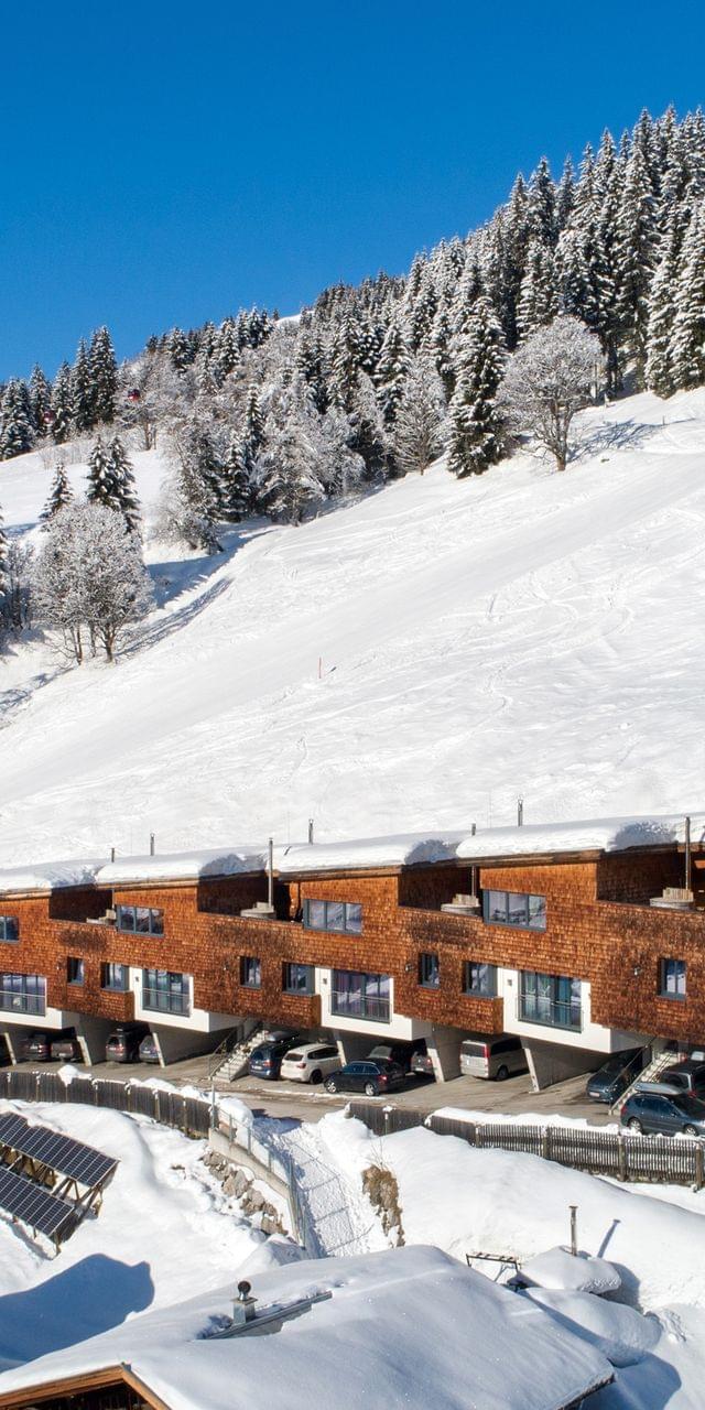 Moderne Holzchalets in verschneitem Alpen-Skigebiet mit bewaldetem Berghang