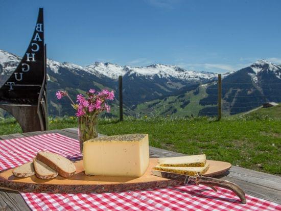 Käse und Brot auf einem Holzbrett vor Alpenpanorama und Frühlingswiese