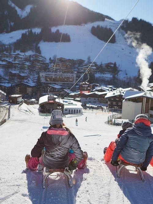 Zwei Familien rodeln bei Sonnenschein einen Hügel zu einem verschneiten Alpendorf hinunter