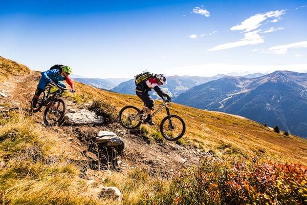 Zwei Mountainbiker fahren bergab auf einem alpinen Trail unter blauem Himmel