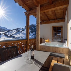 A terrace with a dining table and chairs and a hot tub, overlooking a snowy winter landscape in bright sunshine.