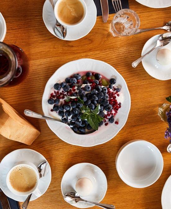 Frühstückstisch mit Joghurt, Beeren, Käse, Schinken, Eiern und Kaffee auf Holzoberfläche