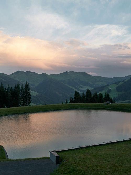 Bergsee mit Spiegelung des Sonnenuntergangs vor Wald und Alpenhügeln