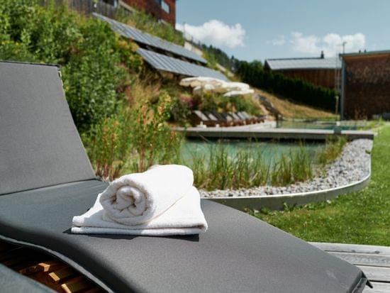 Wellnessliege mit weißem Handtuch neben Naturpool in sonnigem Garten