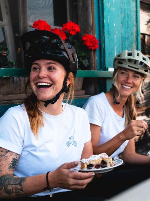 Zwei Frauen mit Fahrradhelmen genießen Kuchen vor einer rustikalen Berghütte