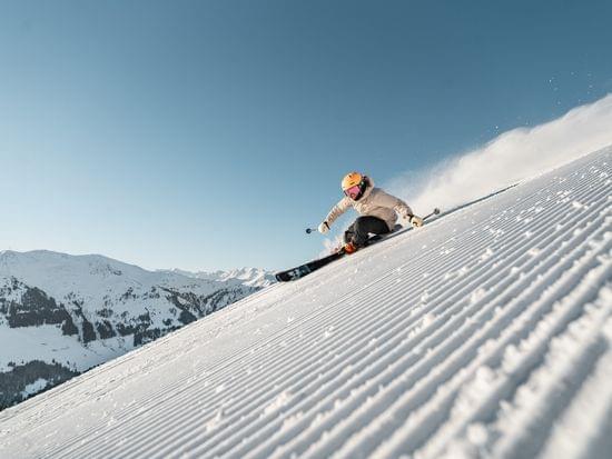 Skier carving downhill on groomed snowy slope with alpine mountains in background