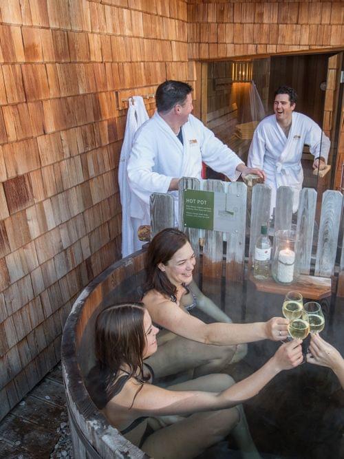 Group of friends enjoying wine and relaxing in a wooden outdoor hot tub at a wellness retreat