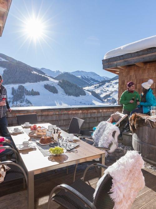 People relaxing on alpine chalet terrace with food and hot tub, snowy mountain view in sunlight