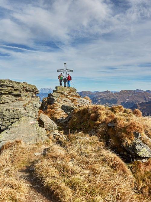 Wanderer am Gipfelkreuz auf felsigem Berggipfel mit Panoramablick auf die Alpen