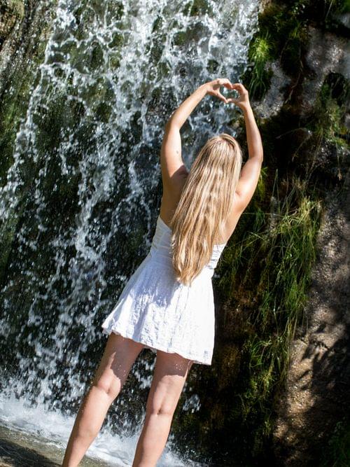 Frau im weißen Kleid formt Herz mit den Händen vor einem Wasserfall
