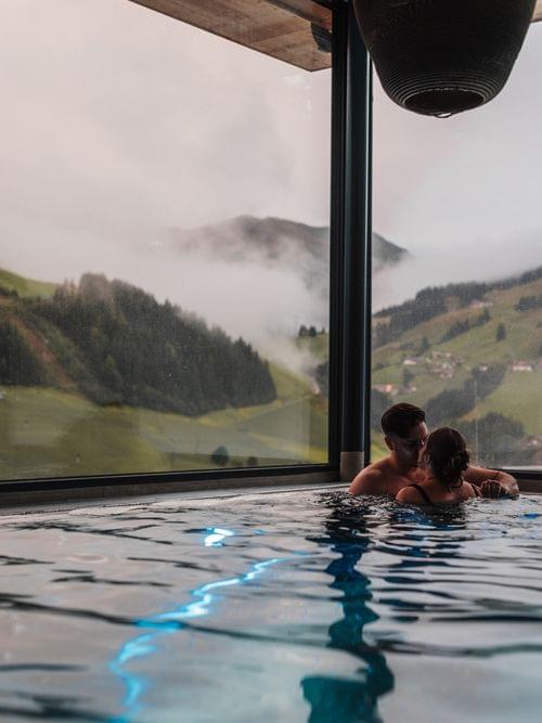 Stimmungsvolles Bild einens Paares, welches im Pool schwimmt und dahinter ein ausblick in die Grüne Berglandschaft 