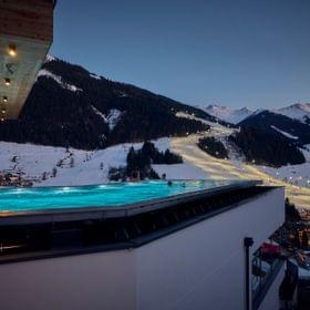 The Bachgut bathhouse infinity pool overlooking the floodlit U-Bahn ski slope, at dusk in a snow-covered winter setting.