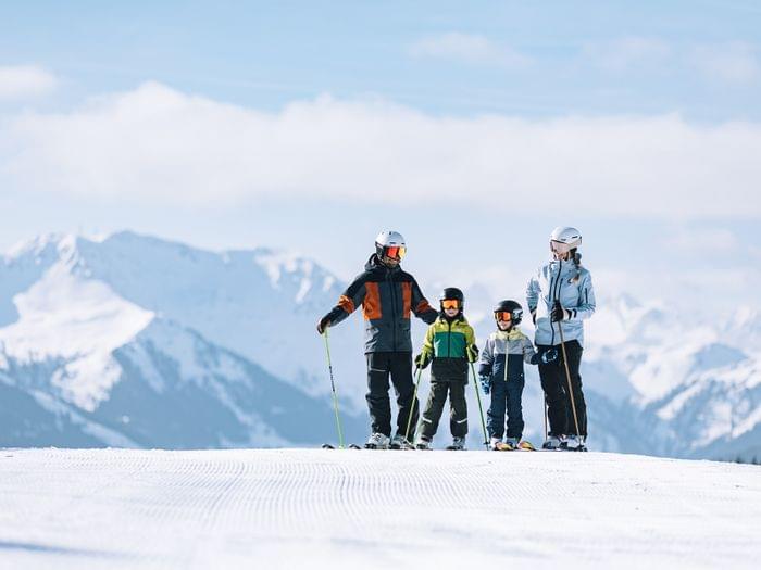 Family skiing on a groomed slope with snowy mountain peaks in the background