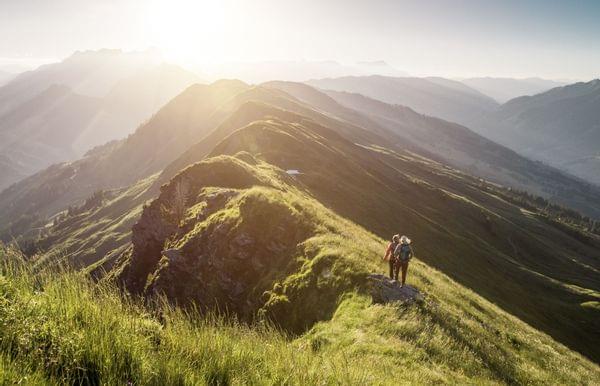 Wanderer auf einem Berggrat bei Sonnenaufgang mit alpiner Landschaft im Hintergrund