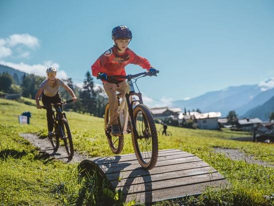 Kind fährt Mountainbike über Holzrampe auf sonnigem Trail mit Bergen im Hintergrund