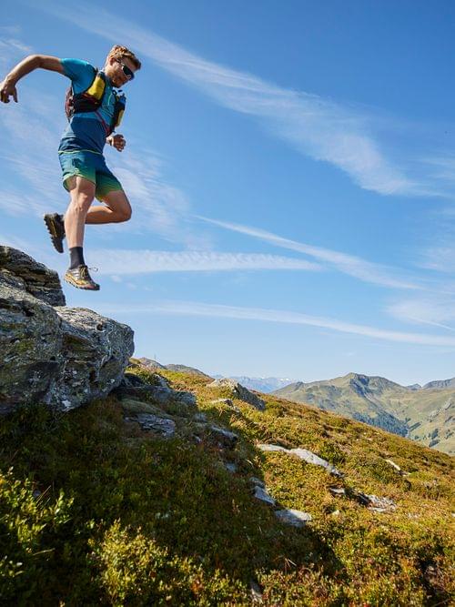 Trail runner jumping off a rock on a sunny mountain trail with scenic alpine view