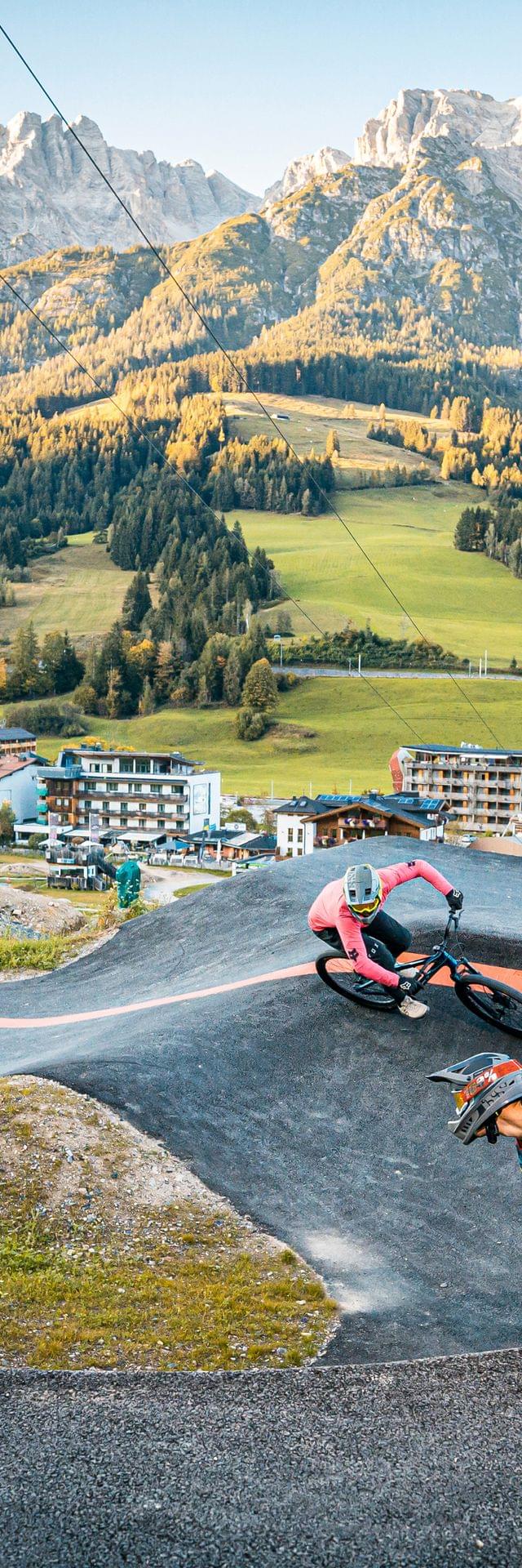 Two mountain bikers riding a pump track in alpine village with scenic mountain backdrop
