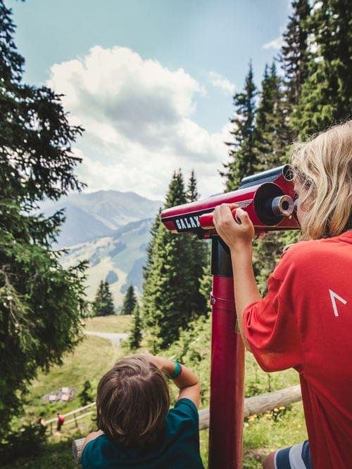 Kinder schauen durch ein rotes Teleskop auf eine Berglandschaft im Wald