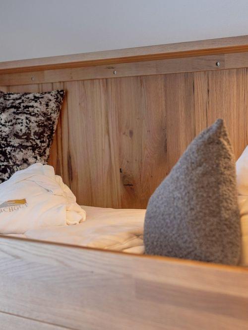 Gemütliches Holzbett im Hotel mit weichen Kissen und gefaltetem Bachgut-Bademantel unter warmem Licht