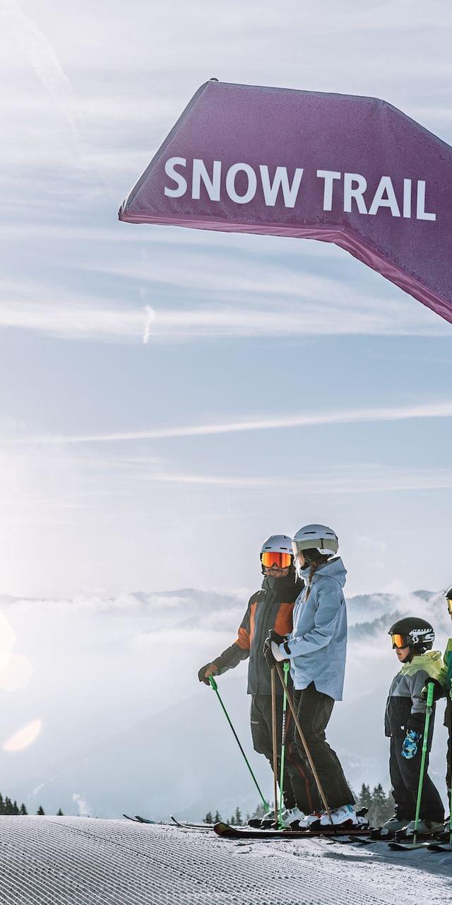 2 Erwachsene mit 2 Kindern beim Skifahren, Sie stehen vor einem Schild mit dem Namen Snow Trail 