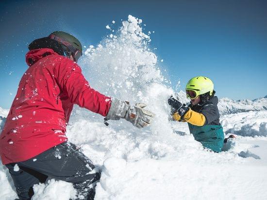 Zwei Personen spielen im frischen Bergschnee unter blauem Himmel in Winterkleidung